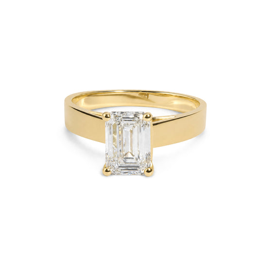 Close-up front angle of a yellow metal ring with a single emerald-shaped white gemstone in a 4-prong setting, shown on a white background.