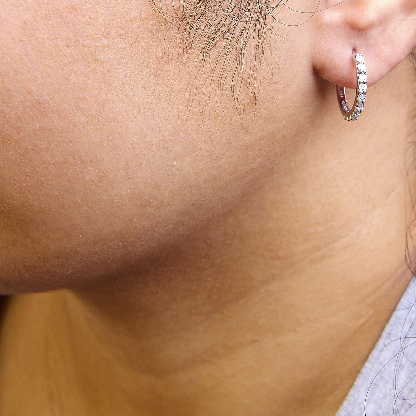 Close-up side view of a white metal hoop earring with round white gemstones in a miracle setting, worn on a model's ear.