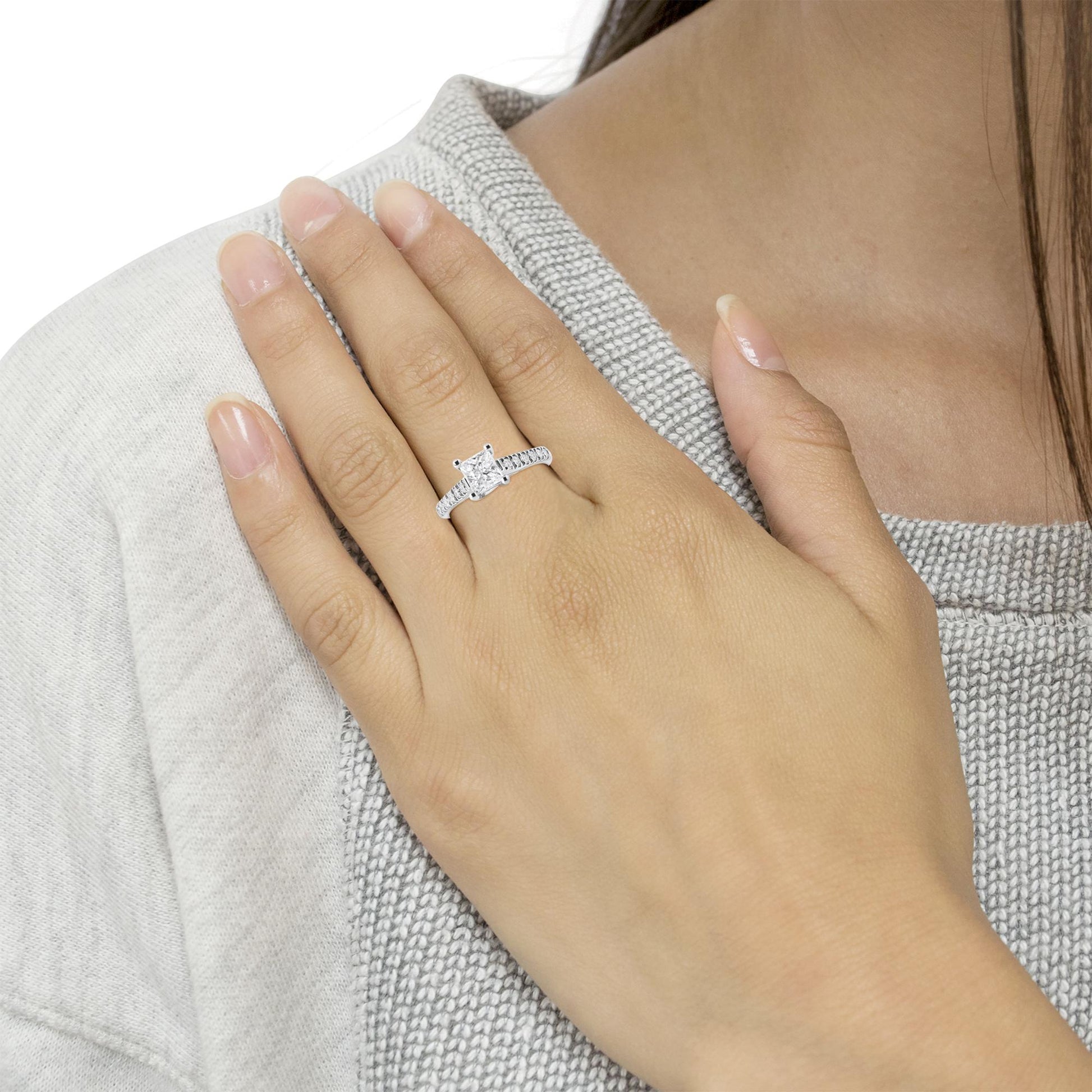 White metal ring with a princess-shaped white gemstone in a four-prong setting and multiple round white gemstones along the band, shown on a model's hand from a top angle.