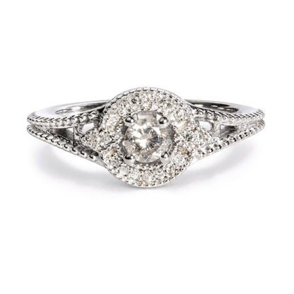 Close-up front view of a white metal ring featuring a central round white gemstone in a prong setting, surrounded by a border of smaller round white gemstones in prong settings, with intricate band detailing.