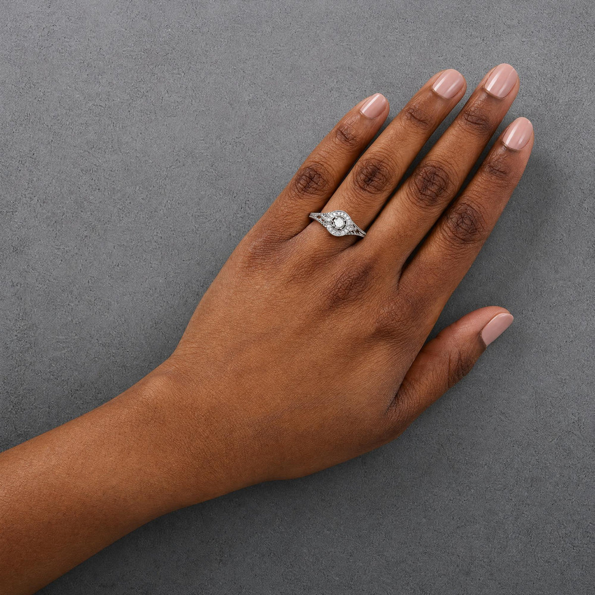 White metal ring with a round white gemstone in prong setting surrounded by smaller round white gemstones in prong settings, shown worn on a model's finger, top-down angle.
