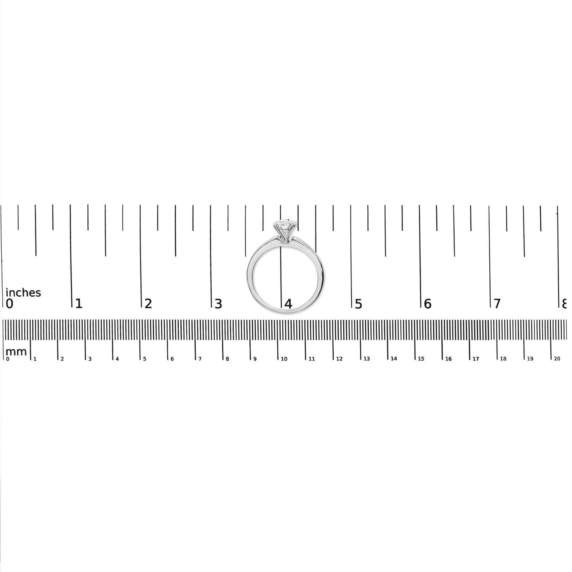 Front-facing view of a white metal ring with a single round white gemstone in a four-prong setting, placed on a ruler for size reference.