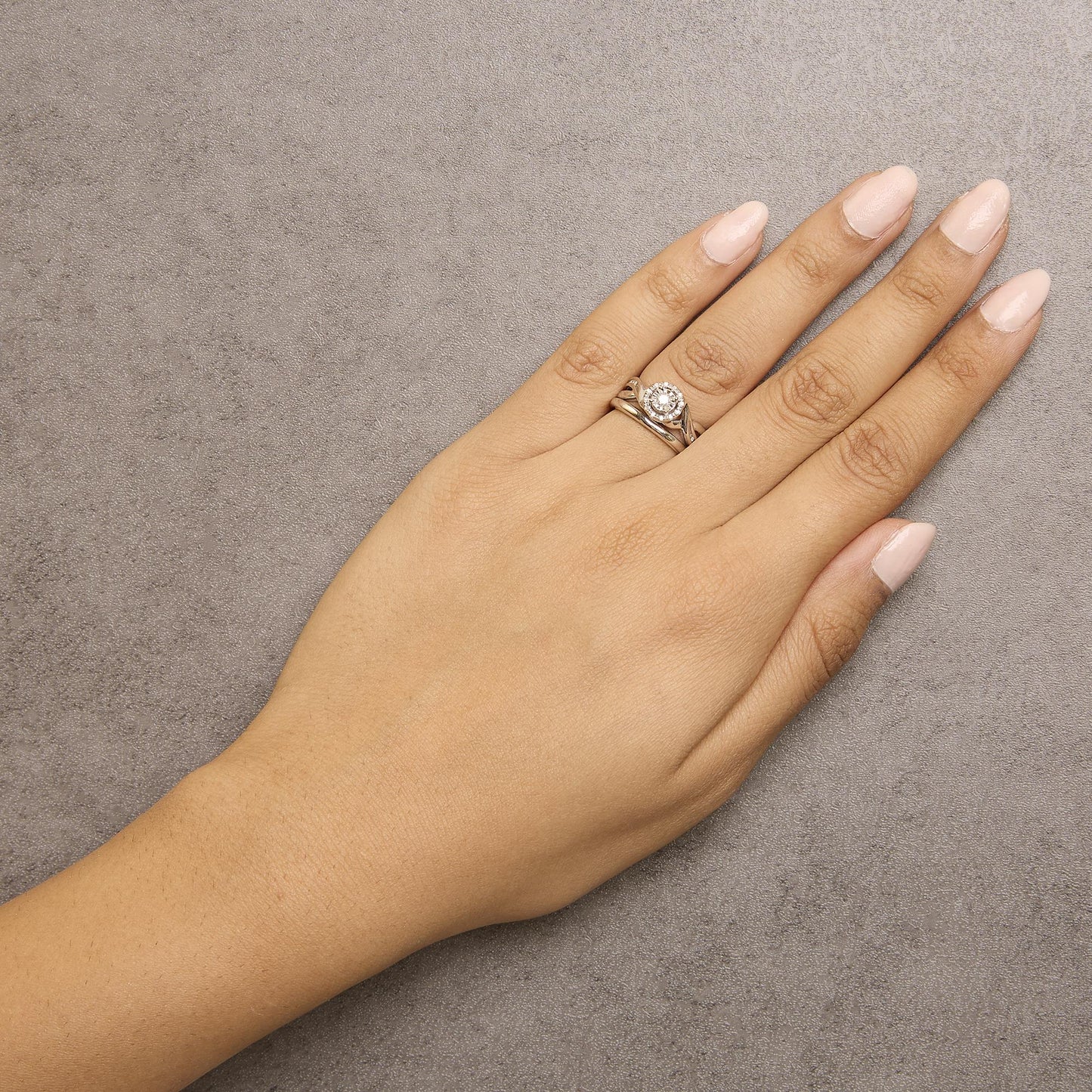White metal ring with a central round white gemstone surrounded by a halo of smaller round white gemstones, worn on a model’s hand, top-down angle.