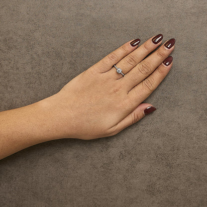 White metal ring with a single round white gemstone in a four-prong setting and multiple smaller round white gemstones in prong settings along the band, shown on a model's hand from a top-down angle.