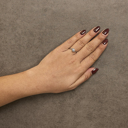 White metal ring with a round white gemstone in a four-prong setting surrounded by smaller round white gemstones in prong settings, worn on a model's hand, shot from above at an angle.