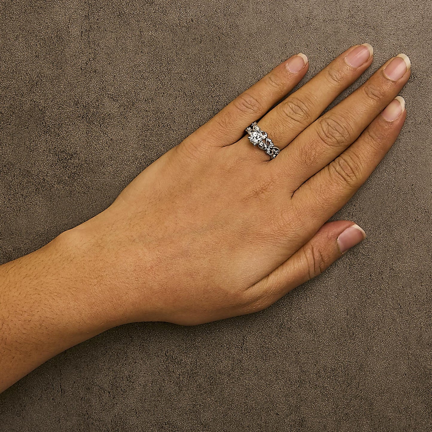Close-up front angle of a white metal ring with two rows of round white gemstones in prong settings, shown worn on a model's hand against a textured brown background.