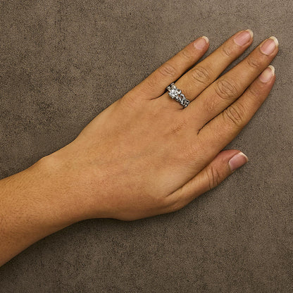 Close-up front angle of a white metal ring with two rows of round white gemstones in prong settings, shown worn on a model's hand against a textured brown background.