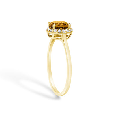 Yellow ring with a round orange gemstone centerpiece in a four-prong setting, surrounded by a halo of round white gemstones in shared prong setting, shown from a side angle on a white background.