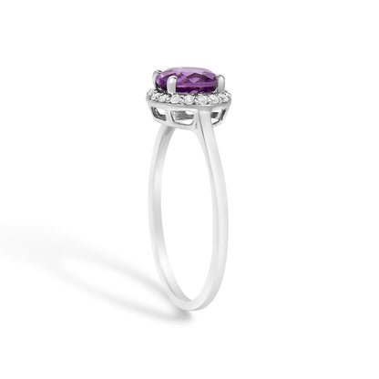 Side angle view of a white metal ring featuring a round purple gemstone in a four-prong setting, surrounded by a halo of round white gemstones in shared prong settings.