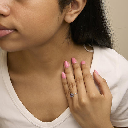 Close-up front angle of a model wearing a white metal ring featuring a round purple gemstone set with four prongs, surrounded by small round white gemstones in a shared prong setting.