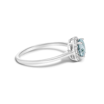 Side view of a white metal ring with a round blue gemstone in a four-prong setting, surrounded by a halo of small round white gemstones in a shared prong setting.