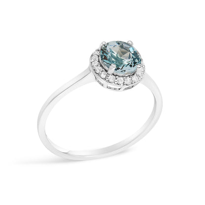 White metal ring with a round blue gemstone held by four prongs at center, surrounded by small round white gemstones in shared prong setting, shown from a slightly elevated side angle on a white background.