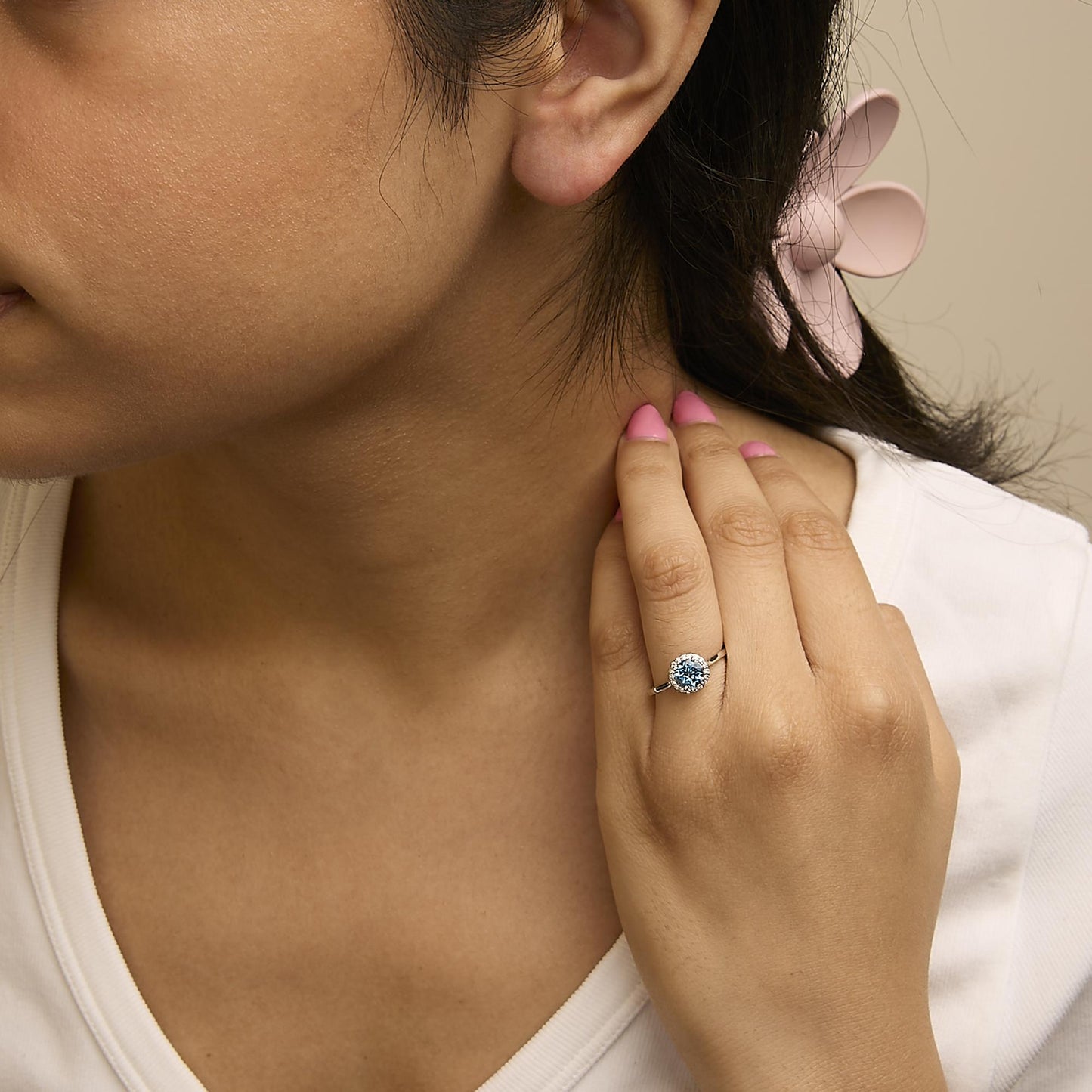 White ring with a round blue gemstone in a 4-prong setting, surrounded by round white gemstones in a shared prong setting, shown worn on a model’s finger in a close-up angled view.