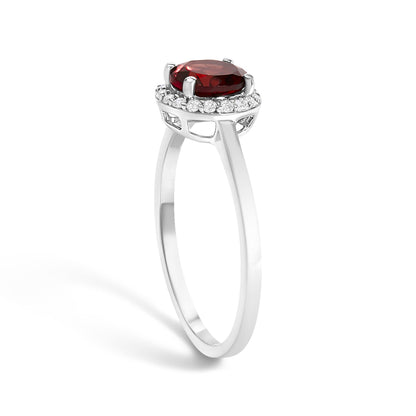 White metal ring with a round red gemstone held by four prongs, surrounded by a circle of small round white gemstones in shared prong setting, shown from a side angle.