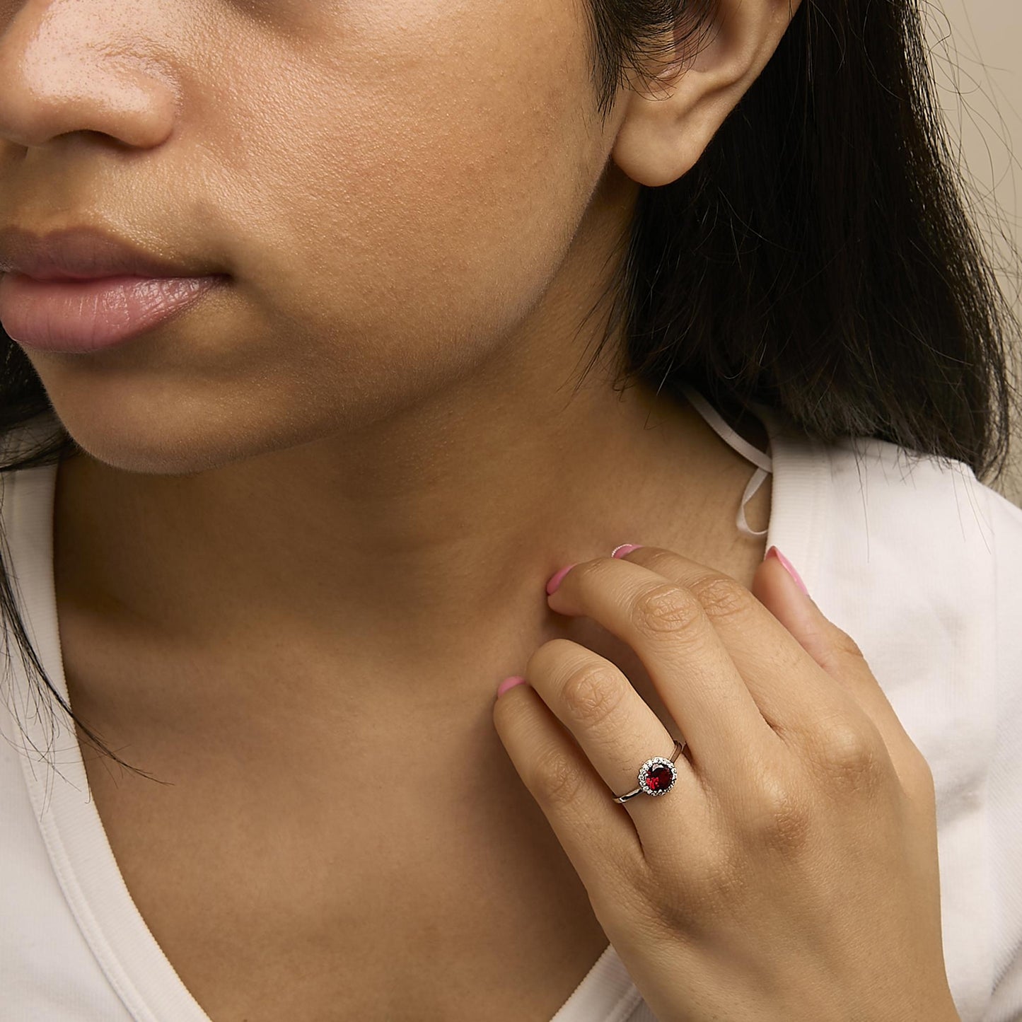 Close-up front angle of a model wearing a white metal ring featuring a round red gemstone center surrounded by a halo of round white gemstones.