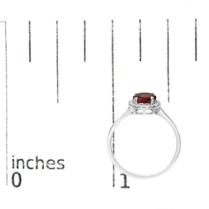 Ring with a white metal band and a round red gemstone in a 4-prong setting, surrounded by multiple small white round gemstones in a shared prong setting; shown on a scale from the side angle.