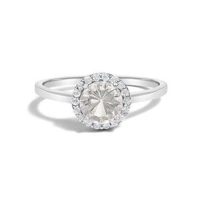 White metal ring with a central round white gemstone in a four-prong setting, surrounded by a halo of smaller round white gemstones in shared prong settings, shown in a front-facing close-up shot.