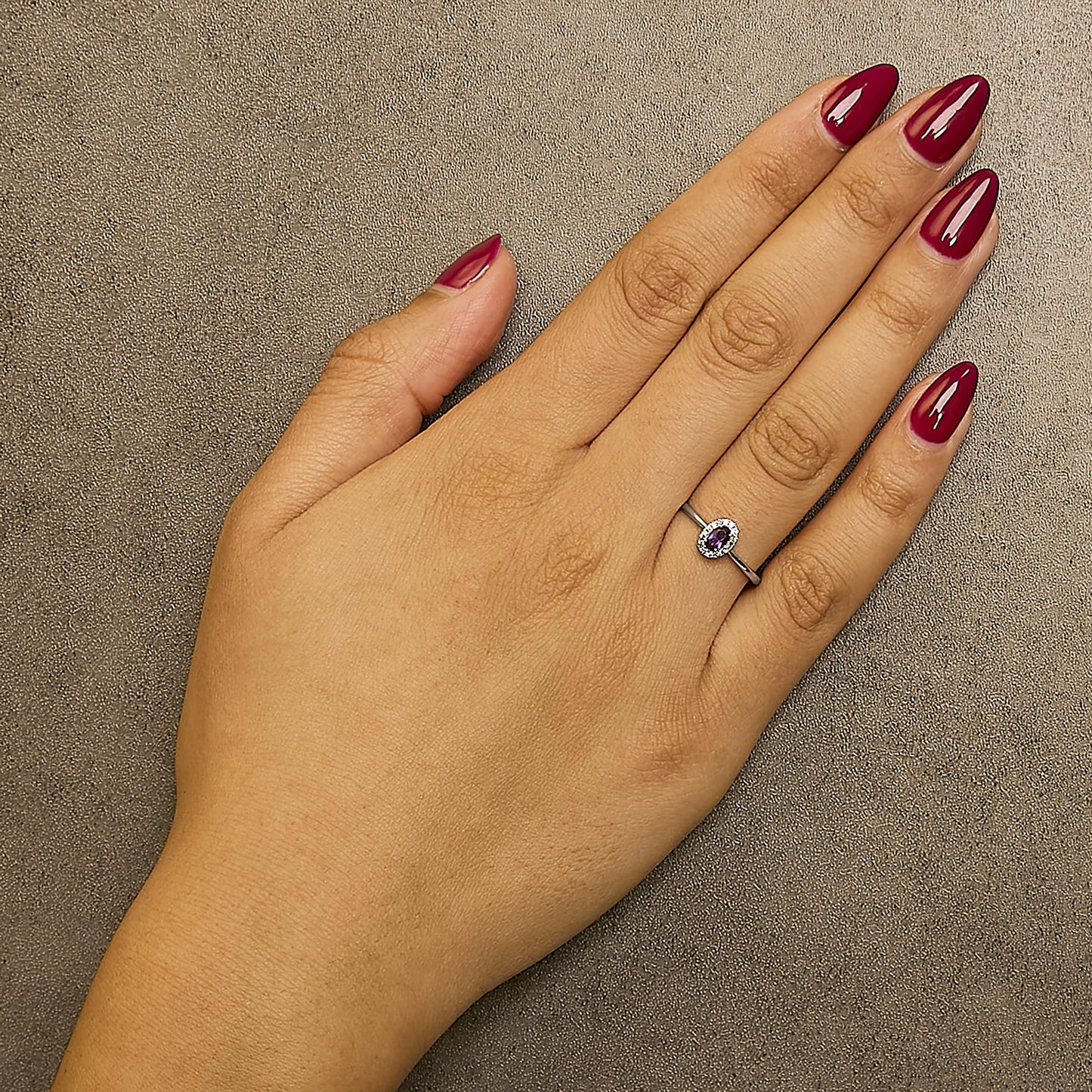 White metal ring with an oval purple gemstone in a prong setting, surrounded by small round white gemstones in a micro-pave setting, shown worn on a model's hand from a top-down angle.