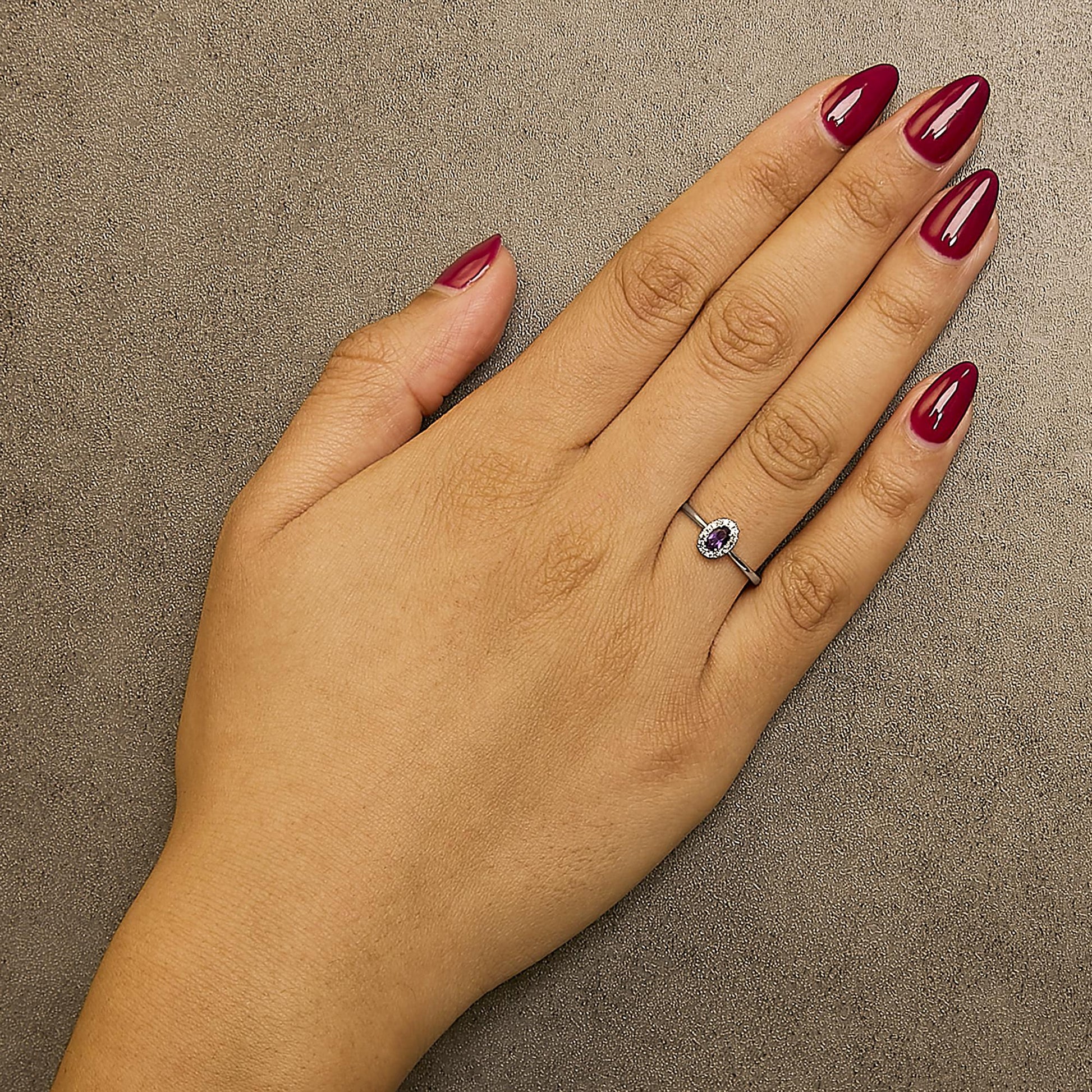 White metal ring with an oval purple gemstone in a prong setting, surrounded by small round white gemstones in a micro-pave setting, shown worn on a model's hand from a top-down angle.