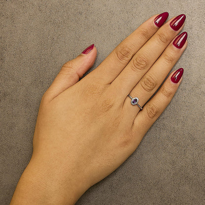White metal ring with an oval purple gemstone in a prong setting, surrounded by small round white gemstones in a micro-pave setting, shown worn on a model's hand from a top-down angle.