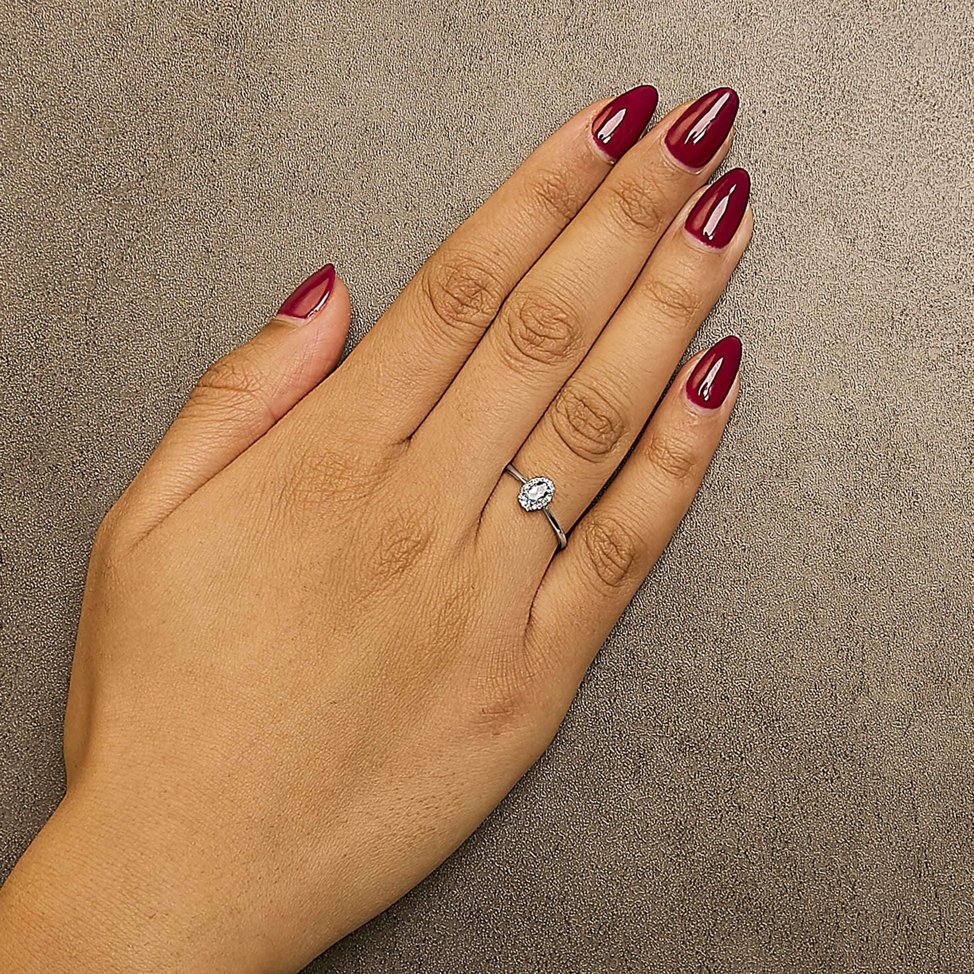 White metal ring with an oval blue gemstone surrounded by small white gemstones in a micro-pave setting, shown worn on a model's hand from a top-down angle.