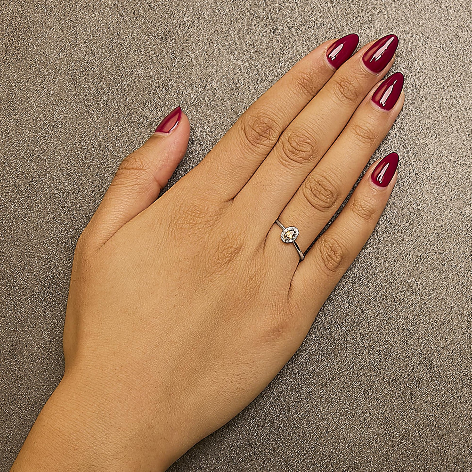White metal ring with an oval orange gemstone surrounded by small white gemstones in a micro-pave setting, shown worn on a model's hand from a top angle.