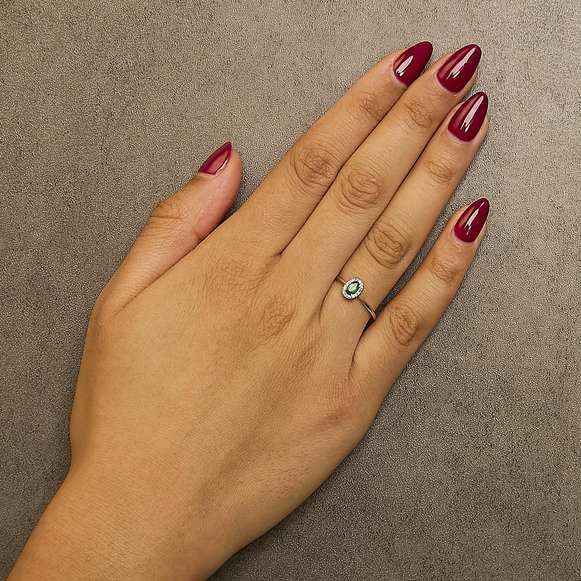 White ring with an oval green gemstone in a prong setting, surrounded by small round white gemstones in a micro-pave setting, shown worn on a model's hand from a top angle.