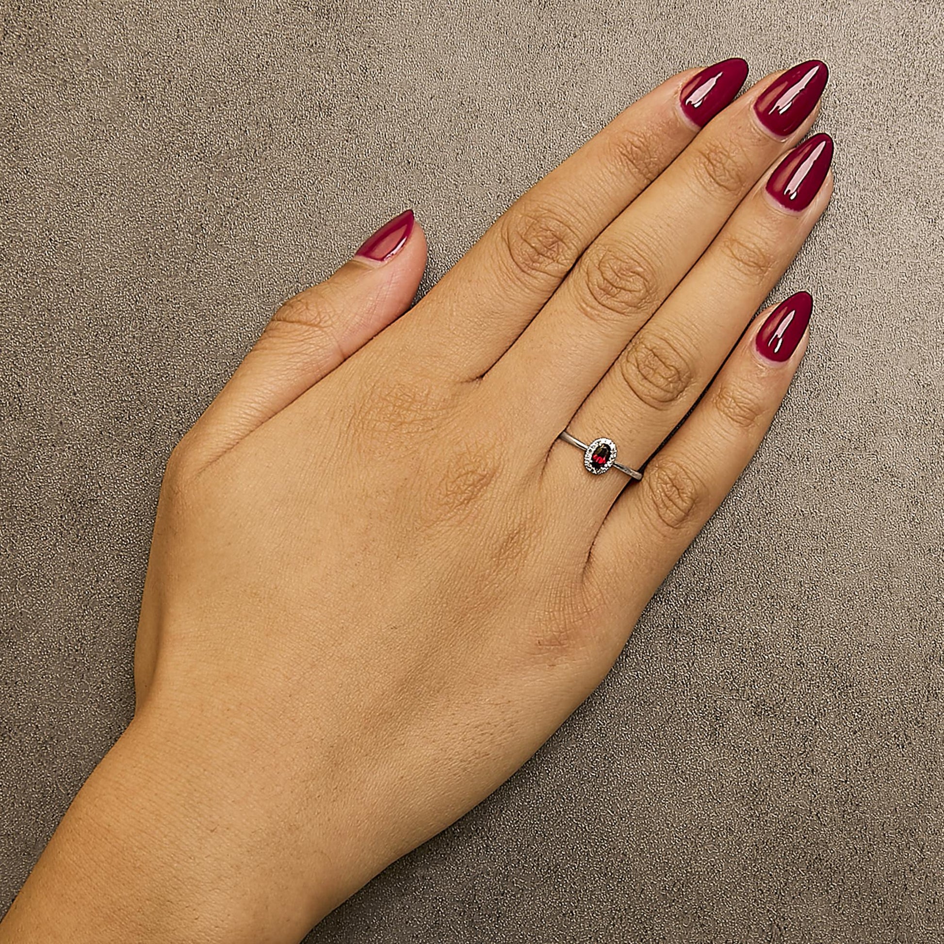 White metal ring with an oval red gemstone in a prong setting, surrounded by small round white gemstones in a micro-pave setting, shown worn on a model's hand from a top-down angle.