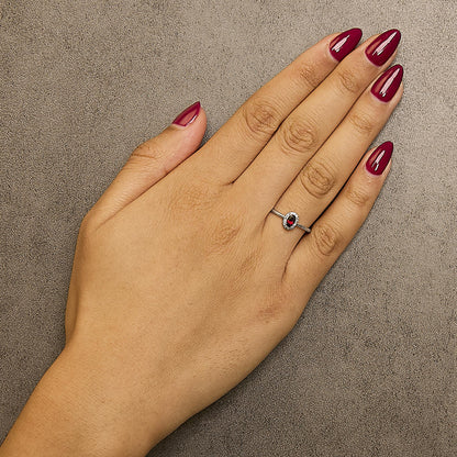 White metal ring with an oval red gemstone in a prong setting, surrounded by small round white gemstones in a micro-pave setting, shown worn on a model's hand from a top-down angle.