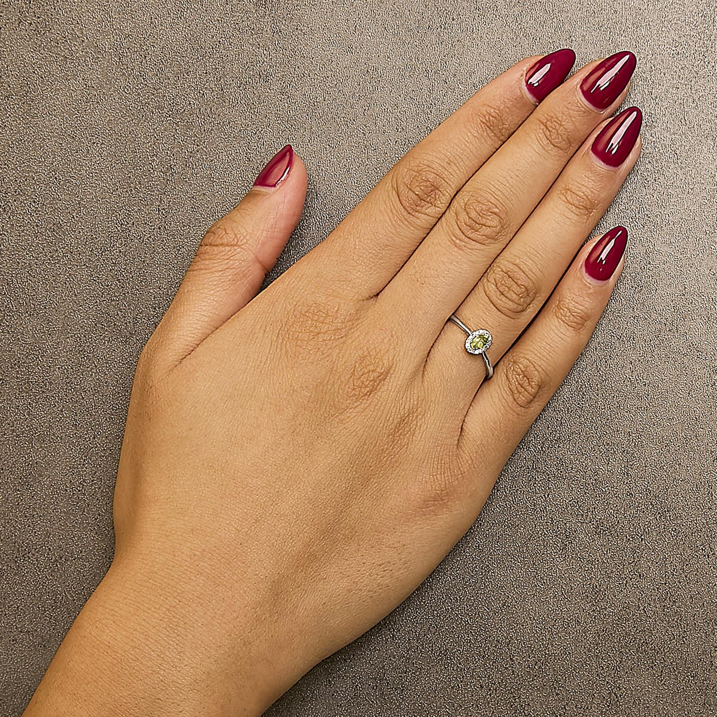 White metal ring with a green oval gemstone surrounded by small white gemstones in micro-pave setting, shown worn on a model's hand from a top-down angle.
