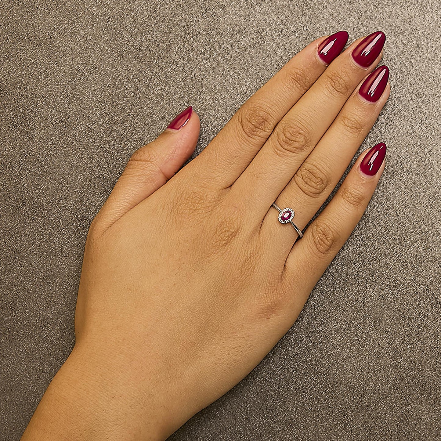Close-up top view of a white metal ring with an oval pink gemstone in a prong setting, surrounded by small white gemstones in a micro-pave setting, worn on a model's finger.