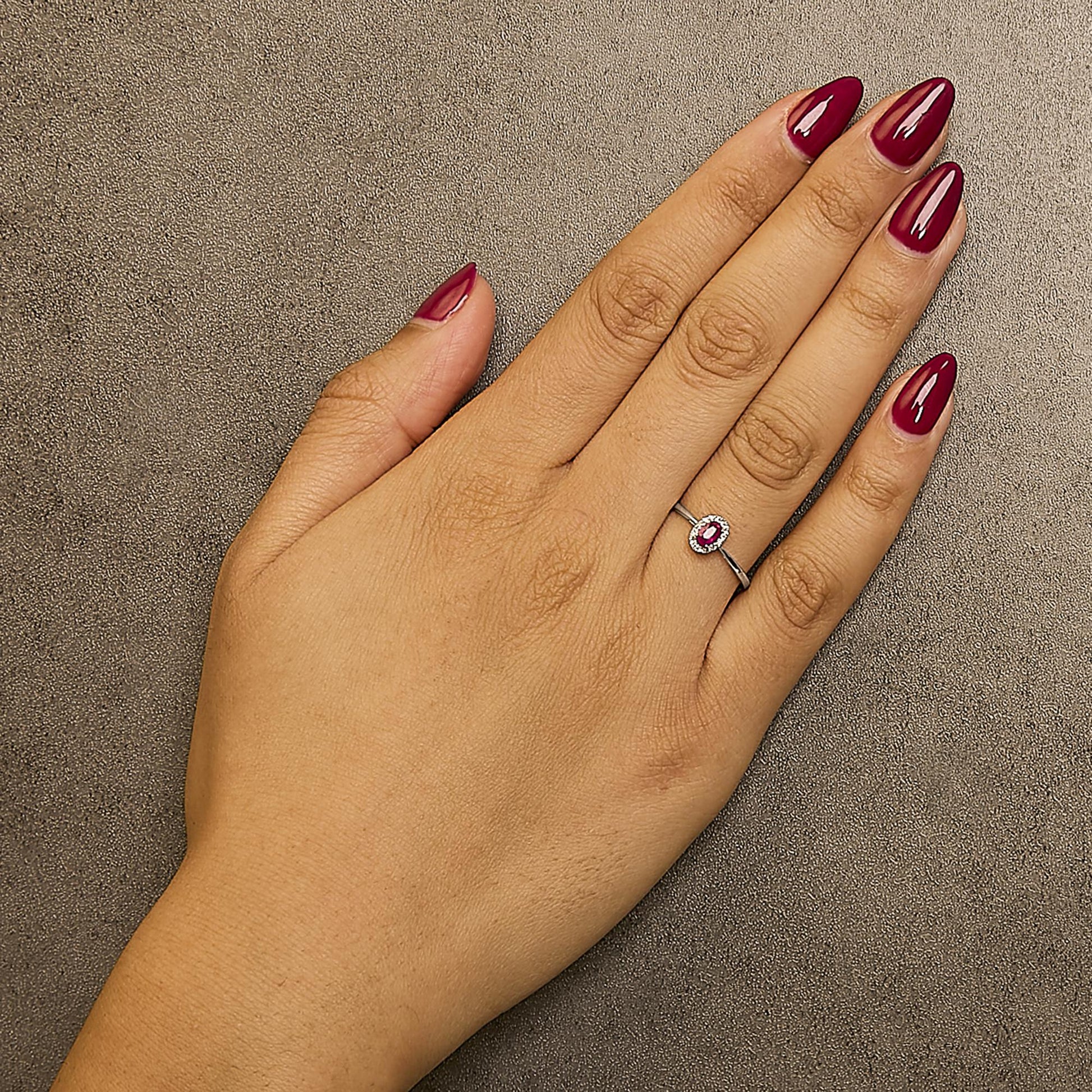 Close-up top view of a white metal ring with an oval pink gemstone in a prong setting, surrounded by small white gemstones in a micro-pave setting, worn on a model's finger.