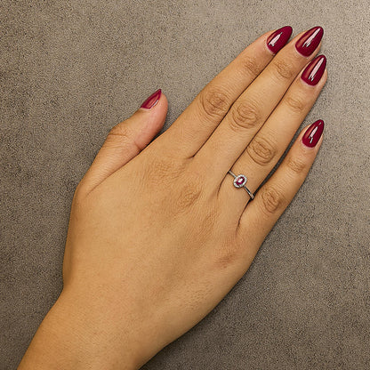 Close-up top view of a white metal ring with an oval pink gemstone in a prong setting, surrounded by small white gemstones in a micro-pave setting, worn on a model's finger.