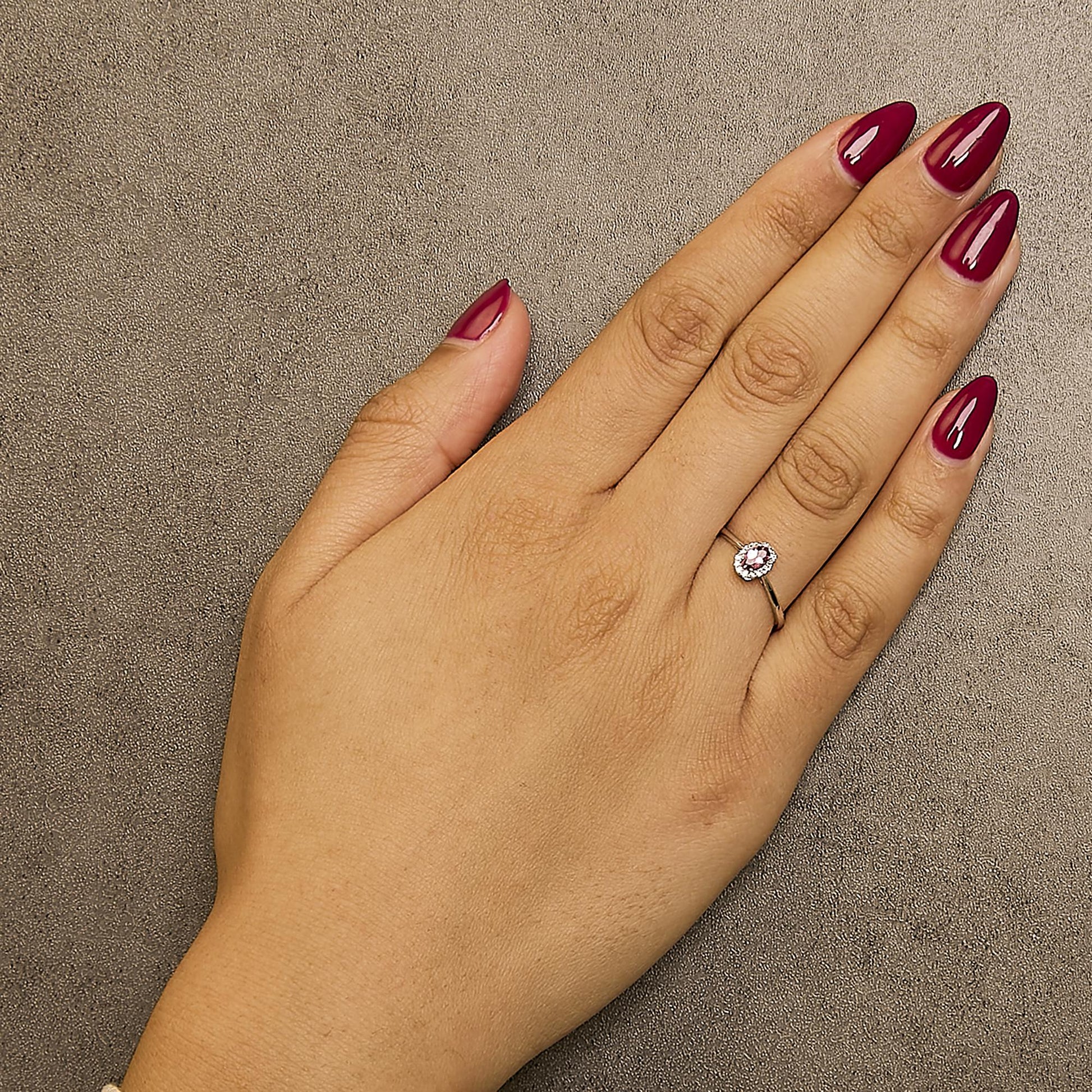 White metal ring with an oval red gemstone surrounded by small white gemstones in a micro-pave setting, shown worn on a model's hand from a top-down angle.