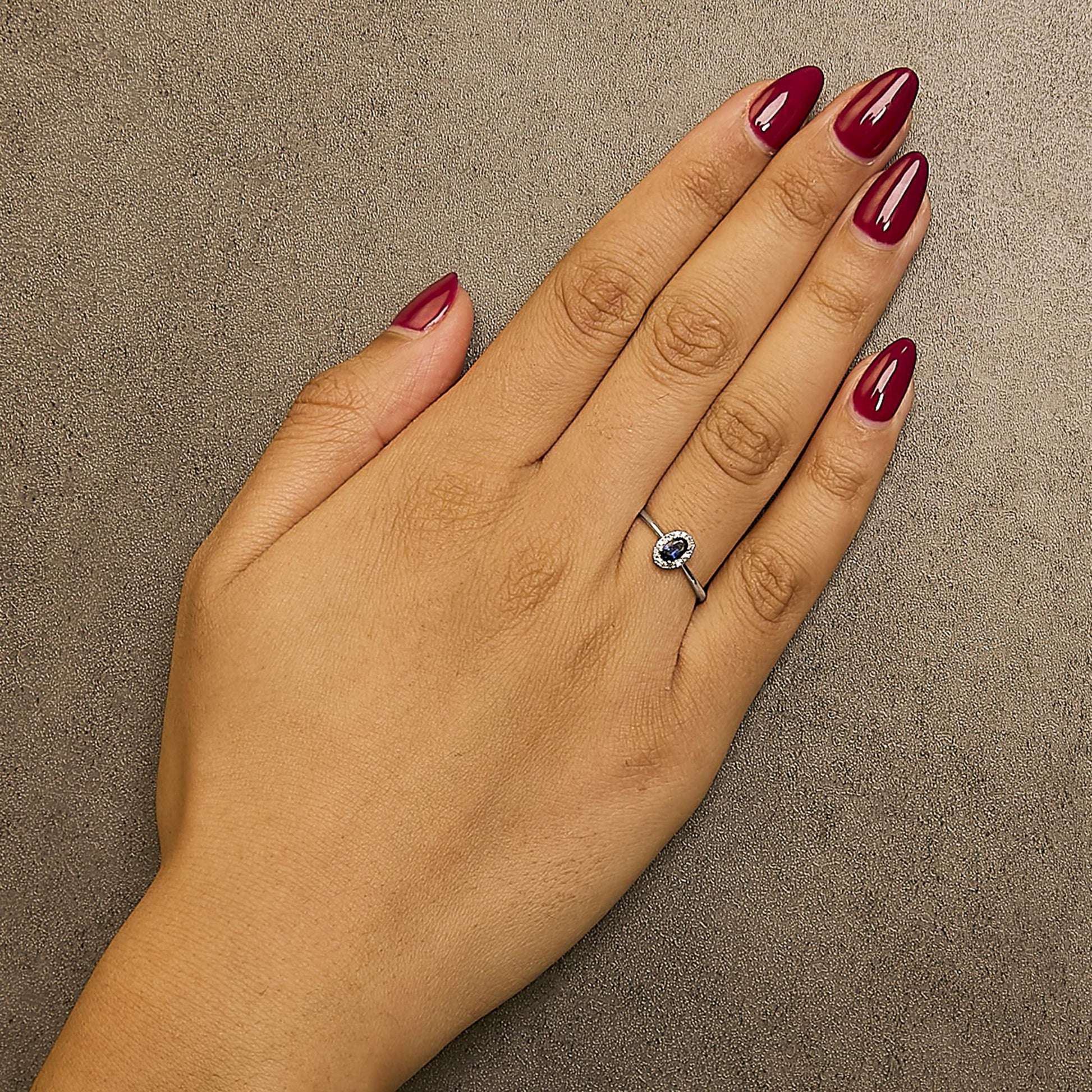 Close-up front angle of a white ring with a blue oval gemstone in the center surrounded by small white round gemstones, shown worn on a model's hand.