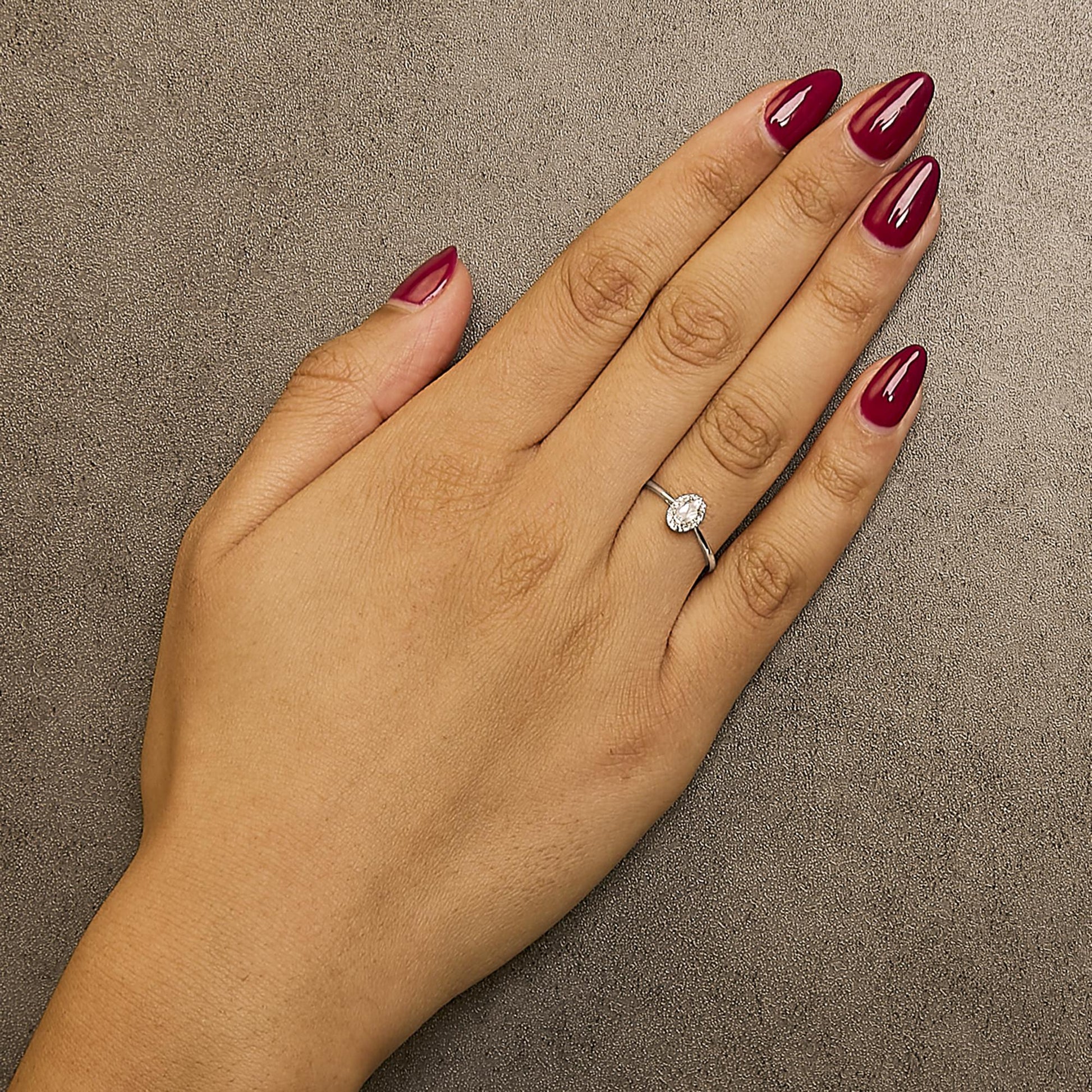 Close-up front angle of a white metal ring with an oval white gemstone in a prong setting, surrounded by small round white gemstones in a micro-pave setting, worn on a model's hand with red polished nails.