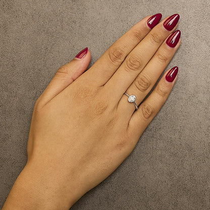 Close-up front angle of a white metal ring with an oval white gemstone in a prong setting, surrounded by small round white gemstones in a micro-pave setting, worn on a model's hand with red polished nails.