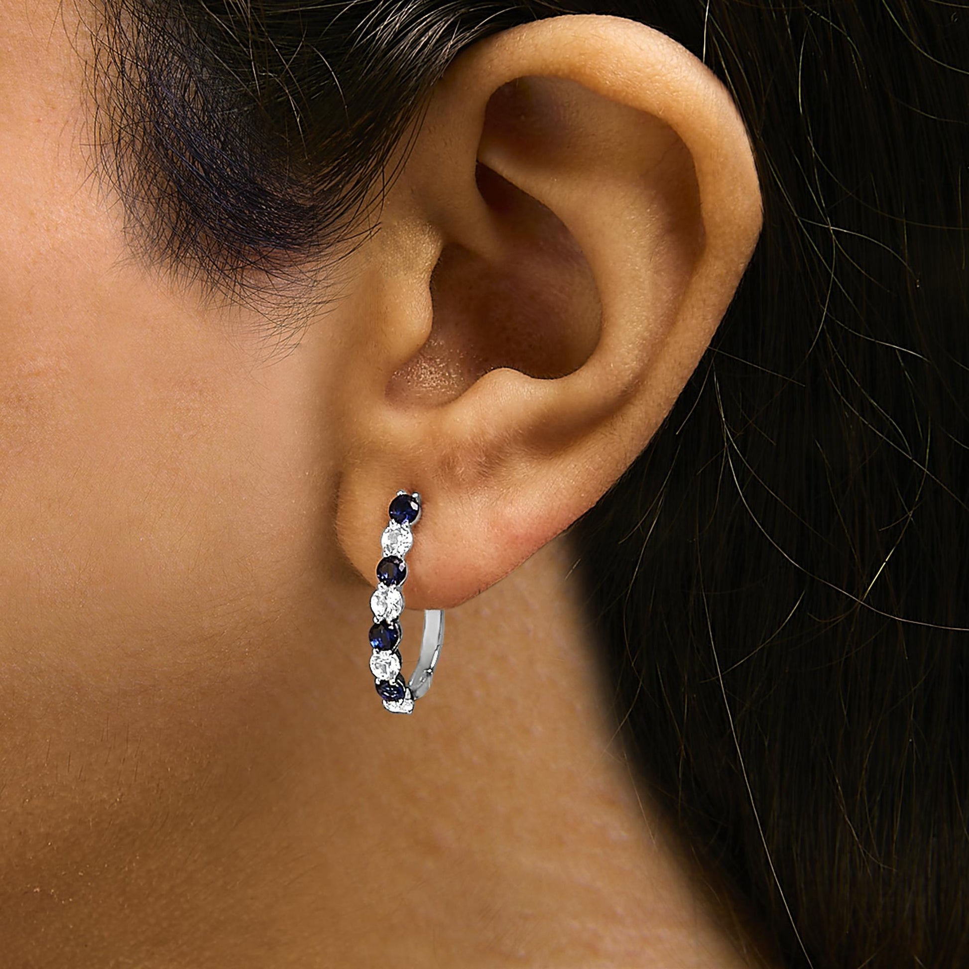 Close-up side view of a white metal hoop earring worn on a model's ear, set with alternating round blue and white gemstones in a shared prong setting.