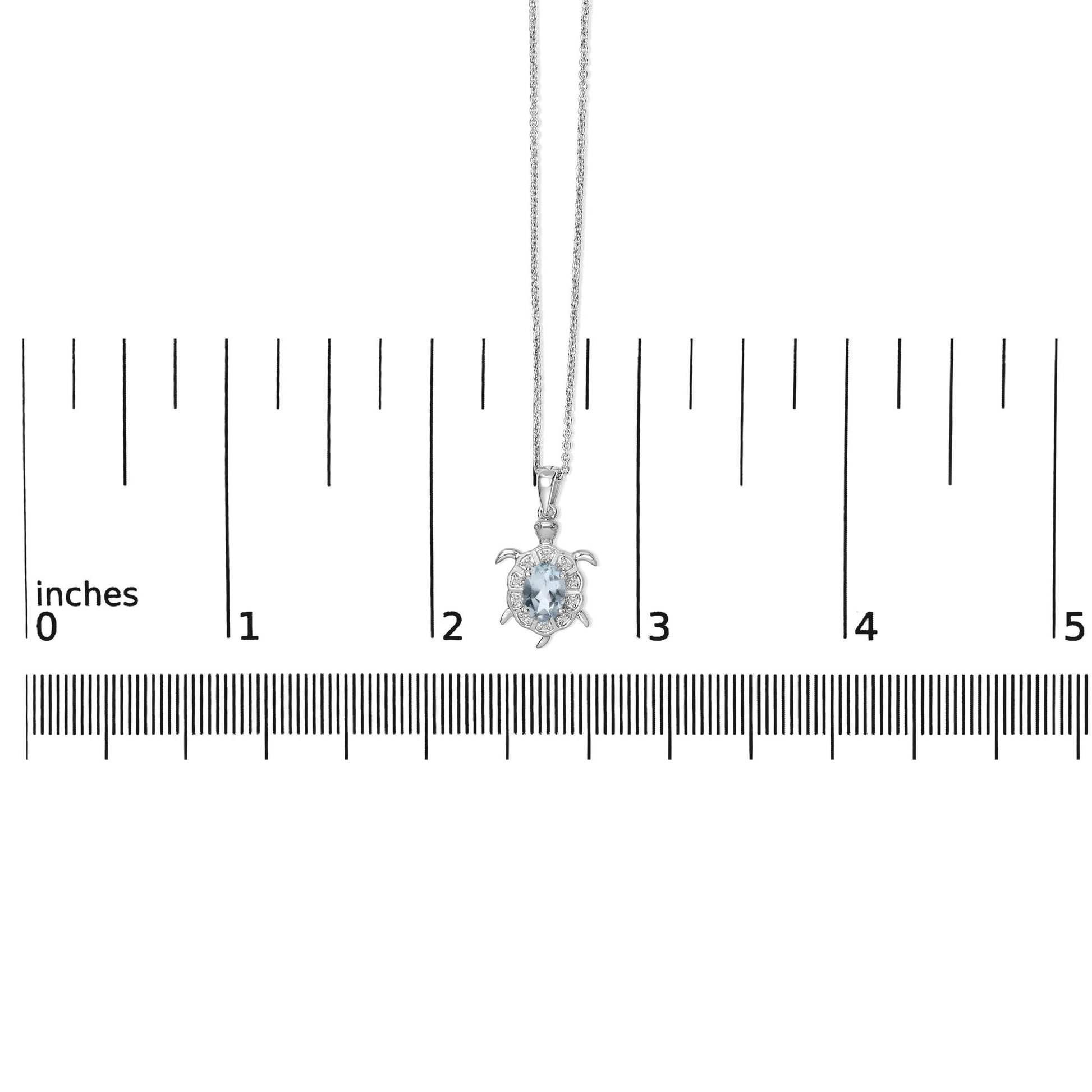 White metal turtle-shaped necklace with a central oval blue gemstone surrounded by small round white gemstones in prong settings, shown on a scale from a front angle.