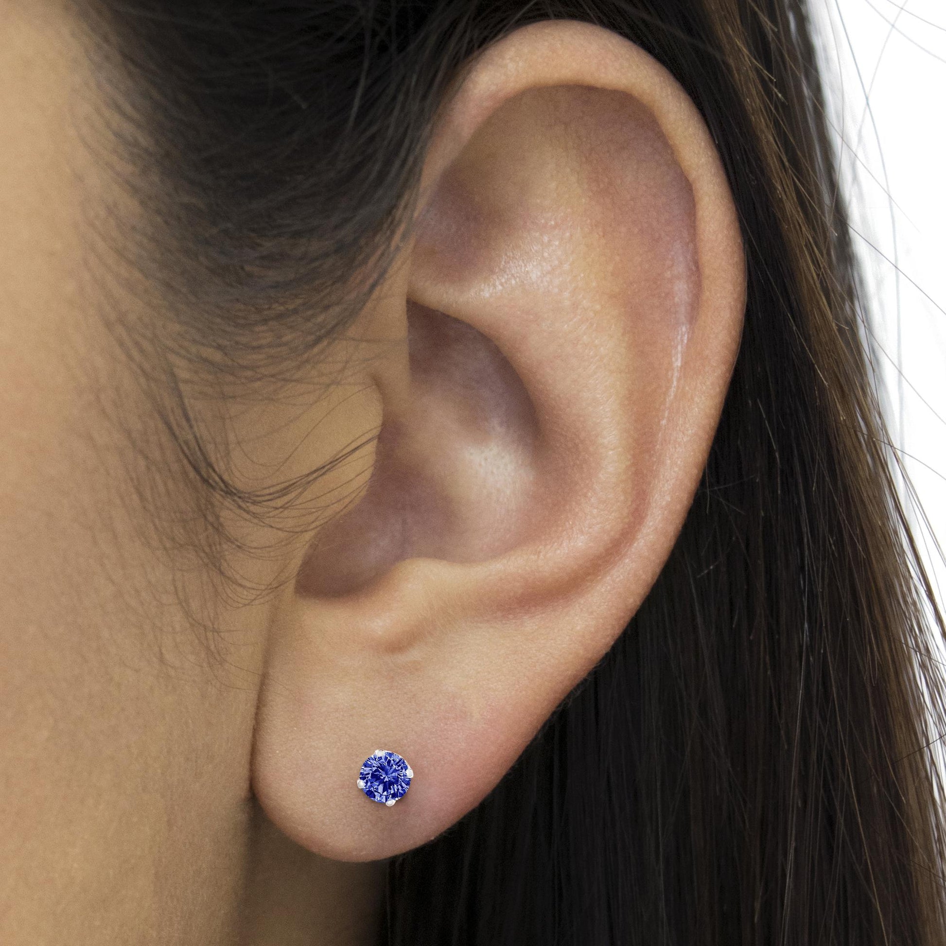 Close-up side view of a white metal stud earring with a round blue gemstone in prong setting worn on a model's ear.