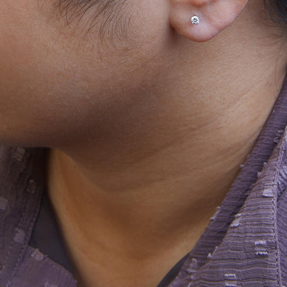 Close-up side view of a white metal stud earring with a round white gemstone in a four-prong setting, worn on a model's ear.