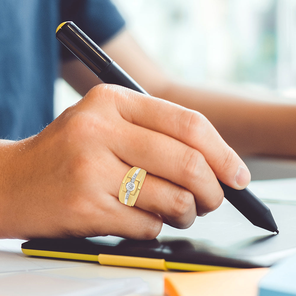 Close-up side angle of a yellow metal ring with a central round white gemstone in a miracle setting, flanked by smaller round white gemstones in prong settings, worn on a person's finger holding a pen.