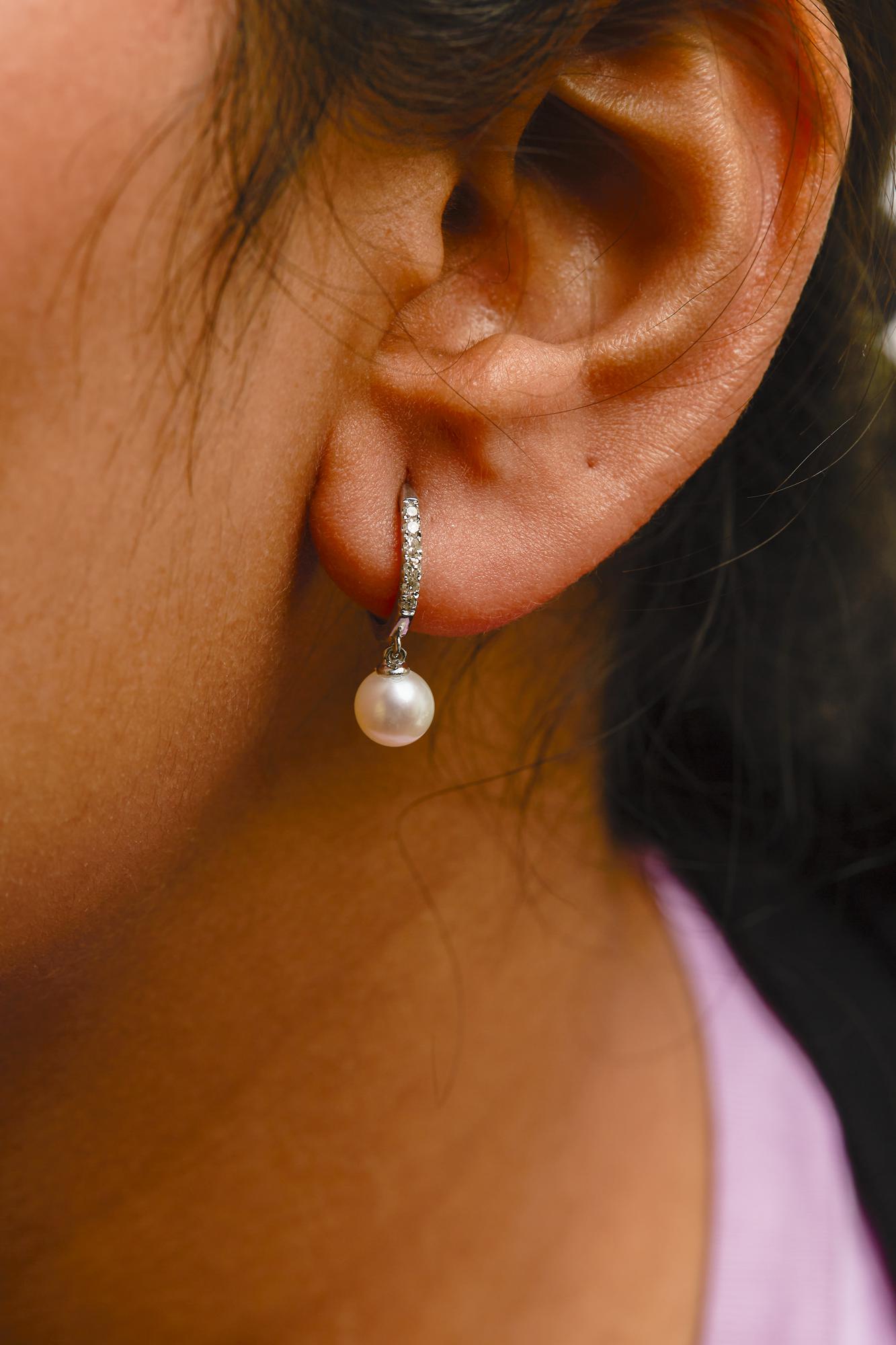 Close-up side view of a white metal hoop earring with pave-set white gemstones and a hanging white round gemstone, worn on a model.