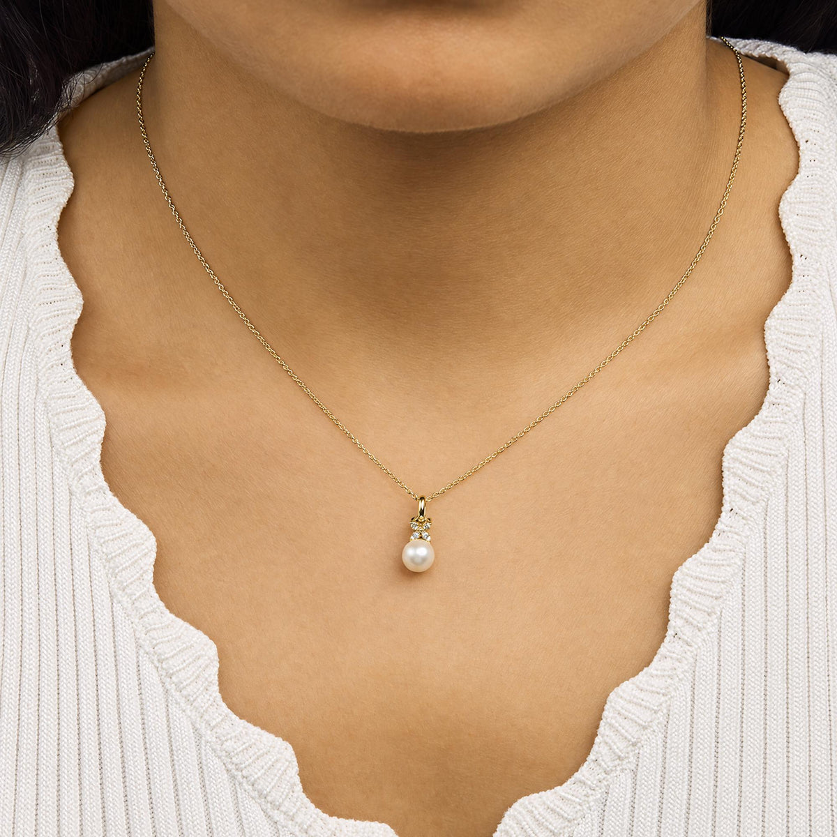 Close-up front view of a model wearing a yellow metal necklace with a pendant featuring a white round gemstone above a pearl, accompanied by several small white round gemstones in a prong setting.