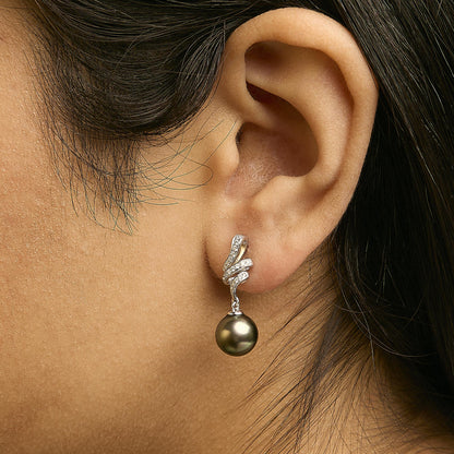 Close-up side view of a model wearing white metal drop earrings featuring a curved micro-pave setting with white round gemstones and a hanging dark round pearl-like gemstone.