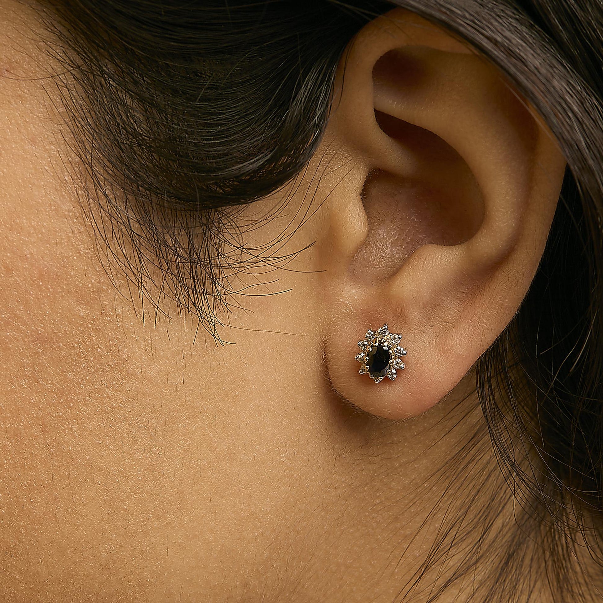 Close-up image of a model wearing a yellow metal earring featuring an oval blue gemstone surrounded by multiple round white gemstones, viewed from the side.