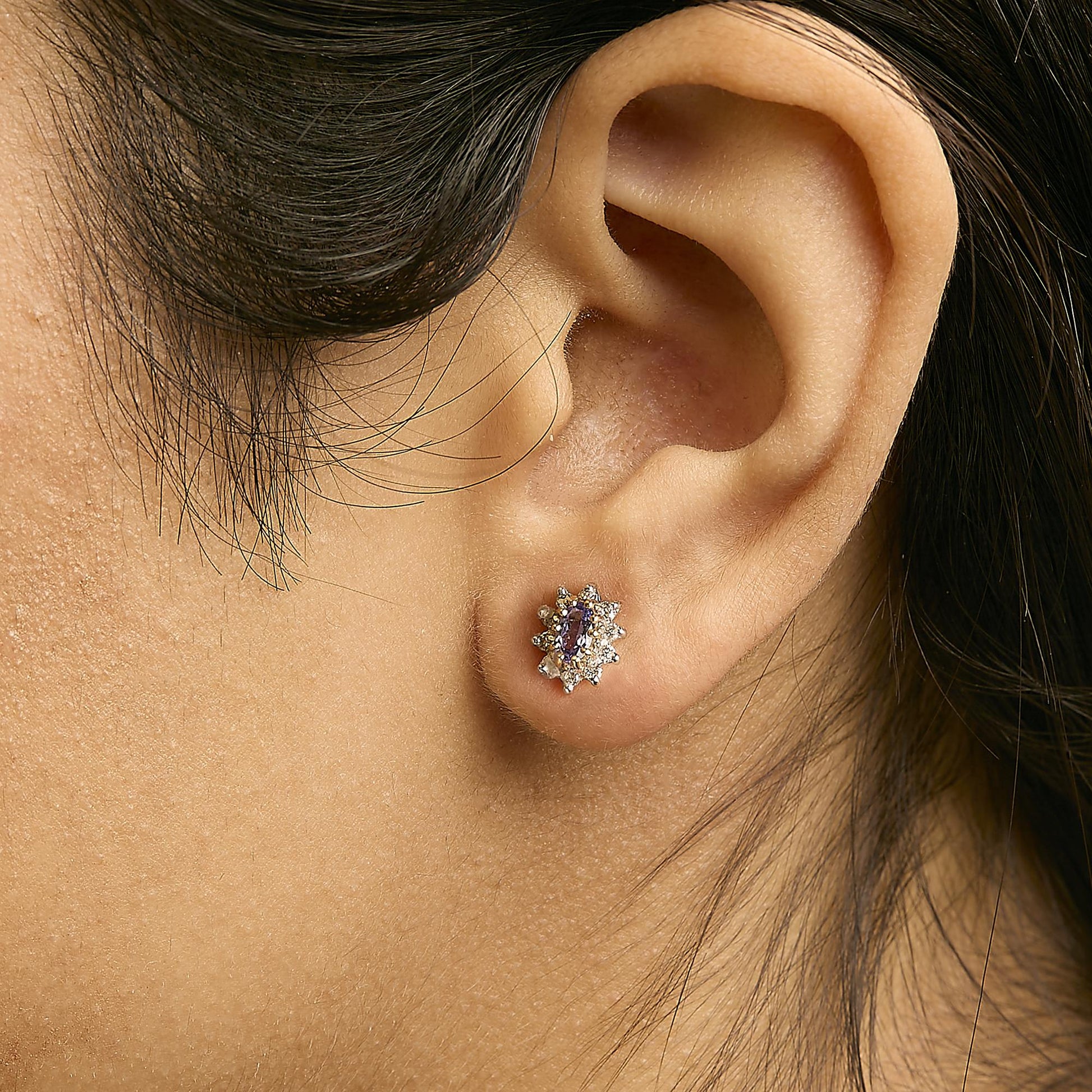 Close-up side view of a yellow metal earring worn on a model's ear, featuring a central oval blue gemstone surrounded by multiple round white gemstones in a prong setting.