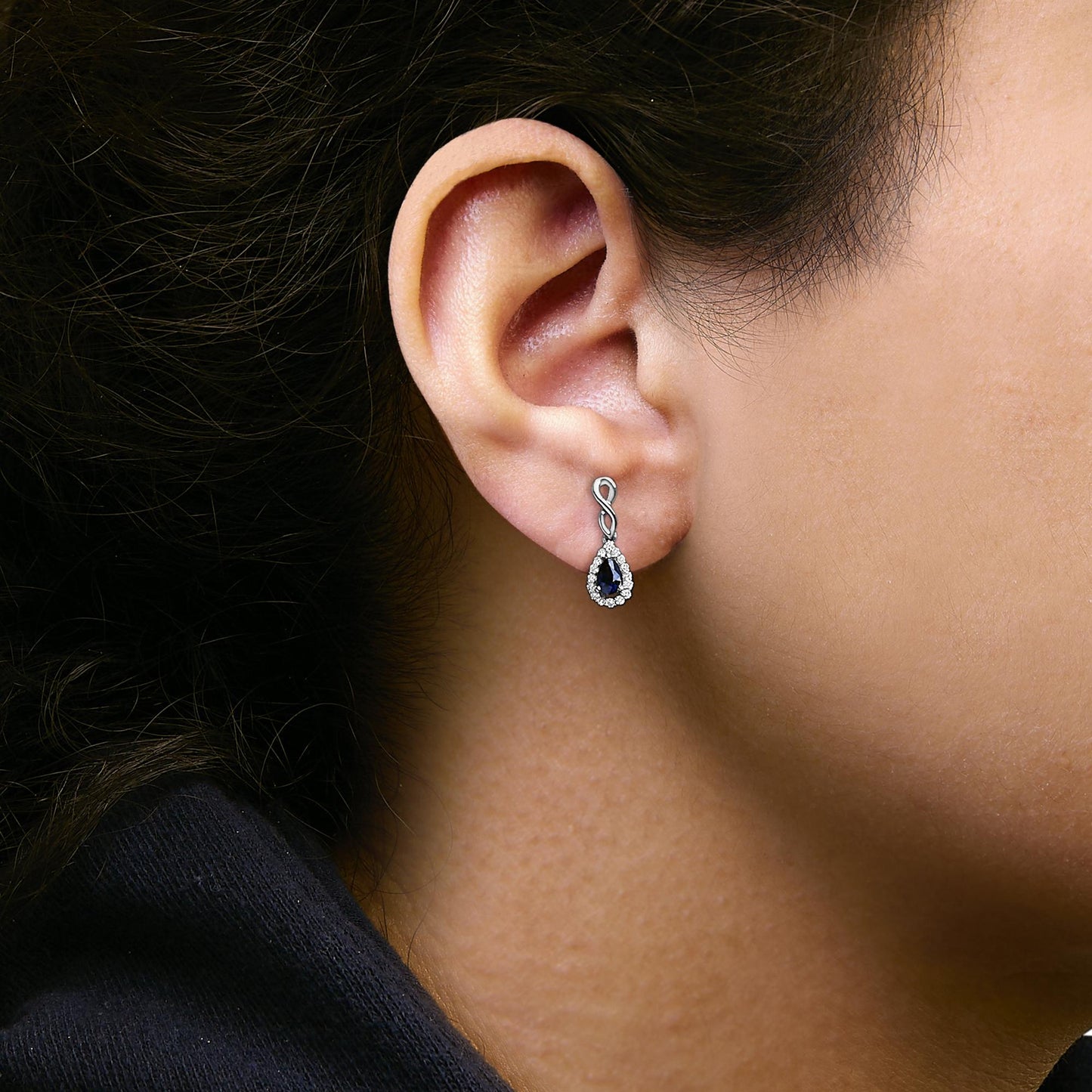 Close-up side view of a model wearing a white metal earring featuring an infinity-shaped design paved with small white gemstones, with a pear-shaped blue gemstone surrounded by white gemstones hanging below.