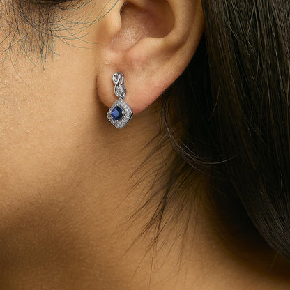 Close-up side view of a model wearing a white metal earring featuring a blue cushion-shaped gemstone surrounded by round white gemstones in a prong setting. The earring has a decorative loop detail connecting to the ear.