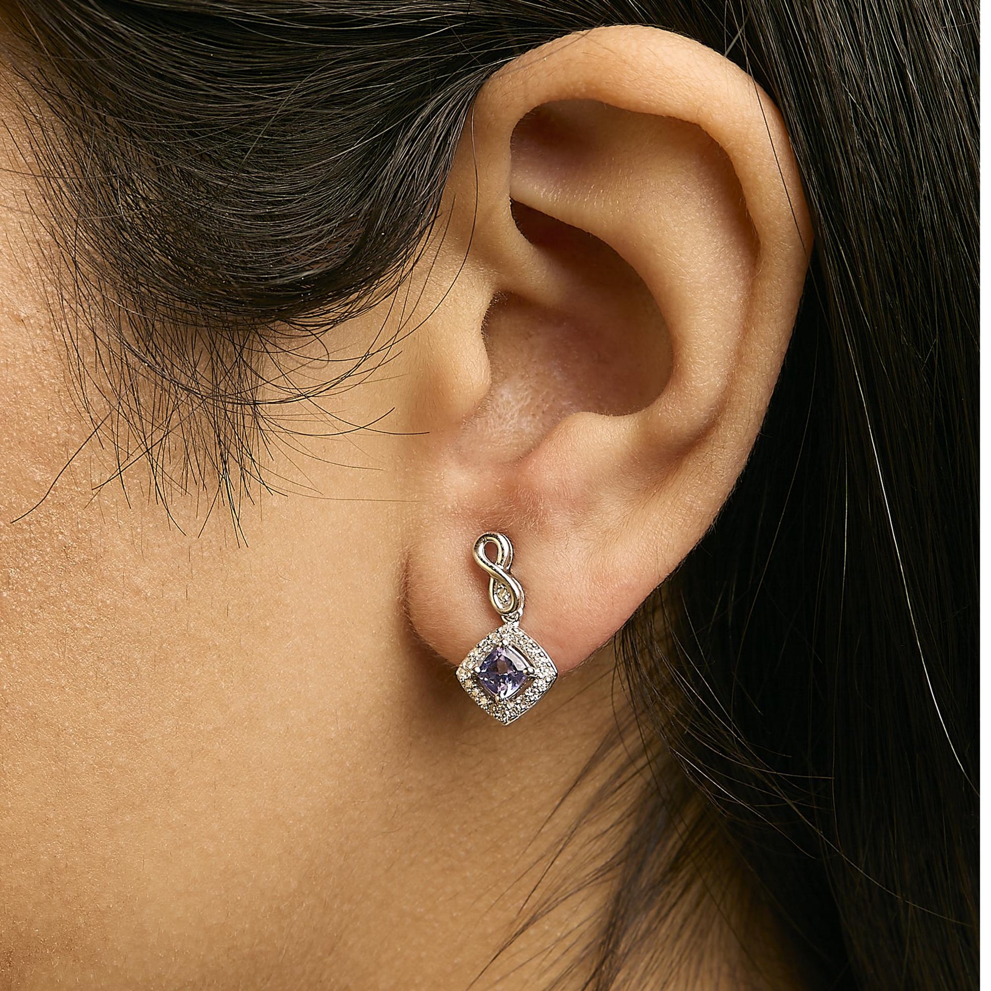 Close-up side view of a model wearing white metal earrings featuring a blue cushion-shaped gemstone surrounded by small white round gemstones, with an infinity loop detail at the top.